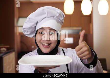 Portrait of Asian female chef looks happy and proud presenting something on her empty white plate, copy space meal menu concept Stock Photo