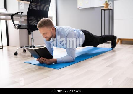 Businessman Reading Notes While Doing Plank Exercise In Office Stock Photo