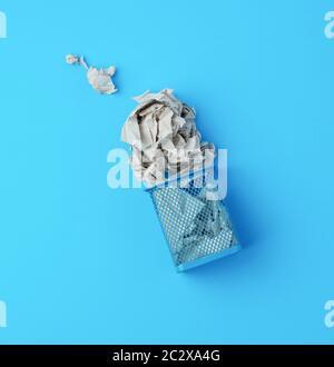 overturned blue metal basket with crumpled pieces of paper, concept of recycling and pollution, blue background Stock Photo