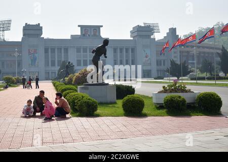 Pyongyang, North Korea - May 1, 2019: A family of locals sits on the sidewalk in front of the Kim Il Sung Stadium Stock Photo