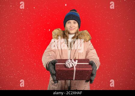 A smiling young woman in winter clothes handsover a red wrapped Christmas present Stock Photo