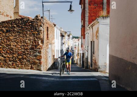 the girl in a helmet and bicycle uniform and white glasses goes with a mountain bike along an old street in Spain and smiling Stock Photo