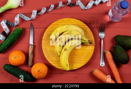 Bananas on wooden plate with avocados, oranges, carrots, scallions, cucumbers and a bottle of water on a red wooden table for diet concept Stock Photo