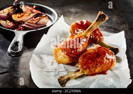 Barbecued spicy chicken legs with hot chili sauce served on crumpled white paper accompanied by a skillet of roast vegetables Stock Photo