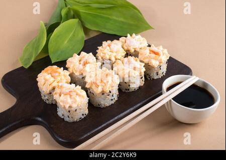 Rolls topped with cream sauce and fried shrimp. Sushi rolls set on dark wooden board. Beige background
