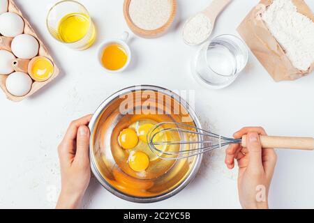 Woman's hands holding whisking eggs in metal bowl next to ingredients for homemade pastry on white table, top view. Stock Photo