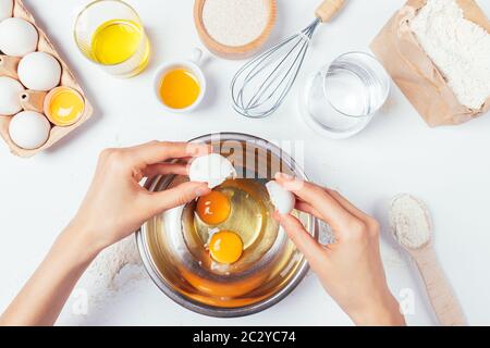 View from above woman's hands crack an egg in bowl to prepare homemade pastry on white kitchen table next to ingredients and utensils. Stock Photo