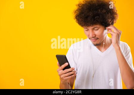Happy asian handsome man with curly hair he smiling enjoying listening to music on earphones using a mobile smartphone isolate on yellow background Stock Photo