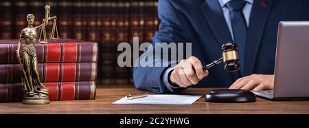 Midsection Of Judge Striking Gavel Near Mallet And Laptop At Desk In Courtroom Stock Photo