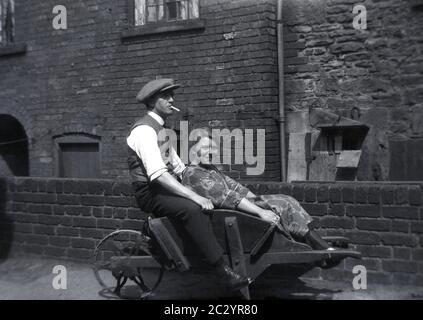 Man in a cloth cap and waistcoat and a cigarette in his mouth, sitting with a young lady on an old wooden framed metal wheelbarrow outside a door covered victorian brick building, common in the industrial areas of Northern England, Sheffield, circa, mid 1930s-1940s. Stock Photo