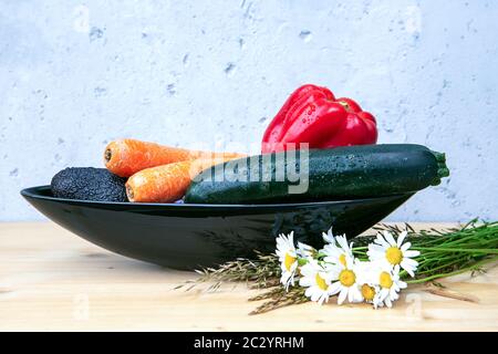 avocado, bell pepper, carrots and cougette in black bowl on wooden table with daisies and grey background Stock Photo