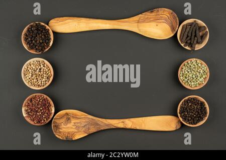 Six small wooden bowls filled with various types of peppercorns and two wooden cooking spoons made of olive wood lie on a black background with copy s Stock Photo