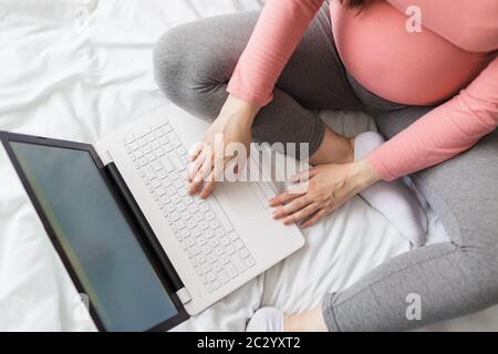 High angle view of young pregnant woman sitting on bed and using laptop. Stock Photo