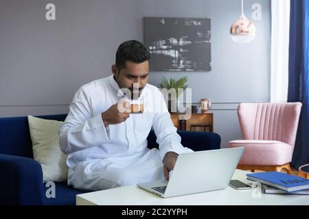 Saudi man drinking tea in living room at home using laptop Stock Photo