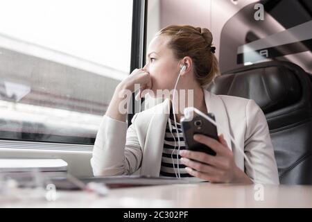 Thoughtful businesswoman looking trough the window, listening to podcast on cellphone using headphone set while traveling by train in business class s Stock Photo