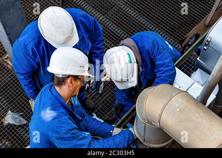 A group of mechanic wearing blue clothes, white helmets and personal protective equipment are assembling a flange at a piping system in Germany Stock Photo