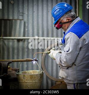 A mechanic with hard hat is doing autogenes cutting of a pipe with a flame of acetylene and oxygen. Sparks are emitted from the material. Stock Photo