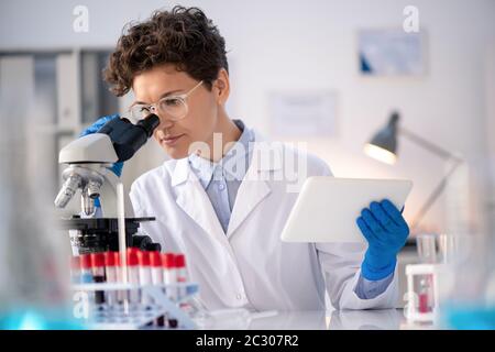 Concentrated laboratory technician using data on tablet while analyzing sample via microscope Stock Photo