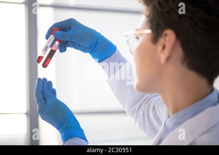 Over shoulder view of female lab specialist in gloves holding test tubes against light while researching blood by coronavirus patient Stock Photo