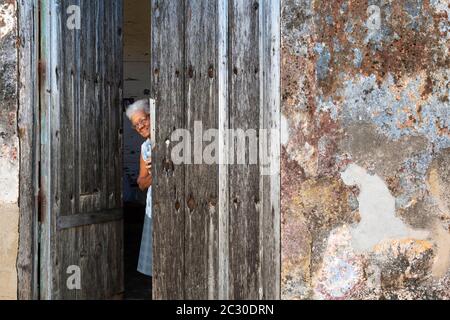Woman at the entrance of her house with a decayed facade of crumbling plaster and a weathered wooden door, Gibara, Cuba Stock Photo