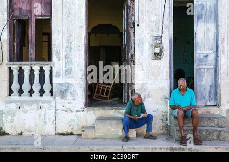 Two men having a chat at the doorsteps of their houses with decayed facades of crumbling plaster and weathered wooden doors, Gibara, Cuba Stock Photo