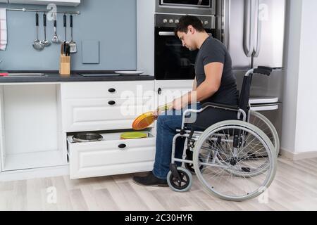 Young Happy Disabled Man Sitting On Wheelchair Arranging Plates In Drawer Stock Photo