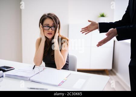 Boss Shouting At Female Employee Sitting At Desk In Office Stock Photo