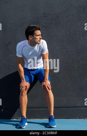 Young running man in blue pants and white shirt resting and recovering on black wall in full sun. Motivation concept, career Stock Photo