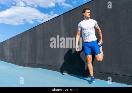 Young man running in blue pants and white shirt resting and recovering on black wall in full sun, doing leg stretches. Motivation concept, career