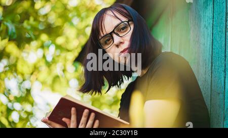 Close up of young woman with glasses reading book in garden. Female resting in nature, enjoying her leisure time. Stock Photo