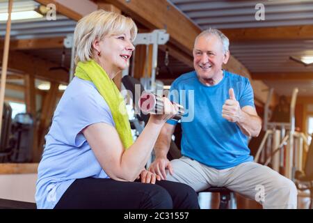 Senior woman in the gym exercising with dumbbells for fitness while her husband fires her up Stock Photo