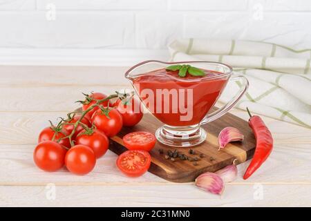 Thick tomato ketchup in a glass gravy boat and ripe cherry tomatoes, garlic and hot pepper on a wood kitchen table. Spices, seasonings and sauces. Stock Photo