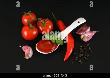 Thick tomato ketchup in a white porcelain spoon,ripe cherry tomatoes, garlic and hot pepper on a black stone surface. Spices, seasonings and sauces. Stock Photo