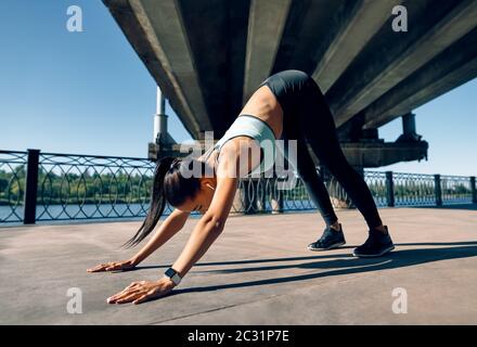 Young sporty woman doing yoga asana downward facing dog under industrial bridge Stock Photo