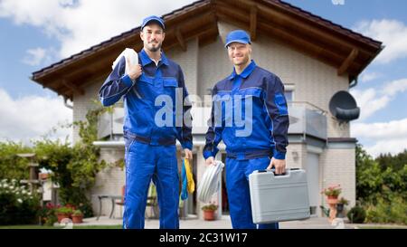 Portrait Of Smiling Two Young Male Electrician Standing In Front Of House With Electrician Kits Stock Photo