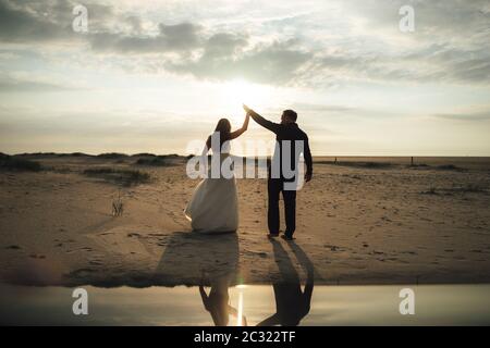 Newlyweds dancing on sandy beach. Bride and groom in evening back-light. Barefoot dance, mirror reflection. Wedding day concept. Stock Photo