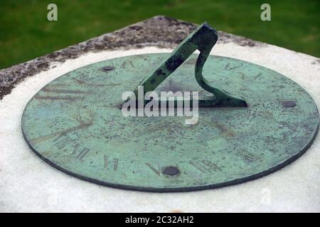 Sundial on stone plinth. Probably made from brass as it has tarnished green. Not enough sun to show the time. Background is lawn grass. Stock Photo