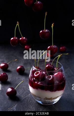 Layered cherry cheesecake in a glass. Dessert made of biscuits, ricotta, cream, cherry jelly and fresh fruits. Dark moody photo. Stock Photo