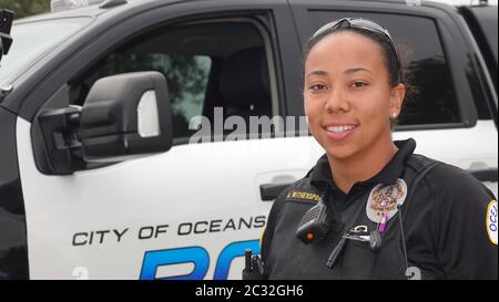 A friendly young female police officer of mixed ethnicity smiles for the camera while standing in front of a police truck. Stock Photo