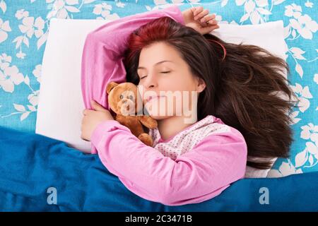 Beautiful young woman in pink pajamas sleeping in bed, hugging a cute plush teddy bear. Stock Photo
