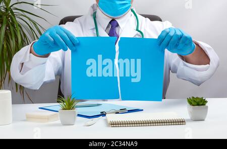 doctor in a white medical coat is sitting at a table in a brown leather chair and holding a torn blue sheet of paper in his hands, place for an inscri Stock Photo