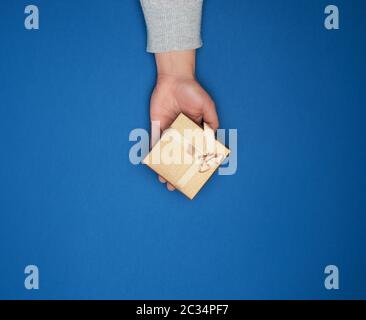 female hand in a gray sweater holds a closed square cardboard gift box with a bow on a blue background, trend colors, holiday backdrop Stock Photo