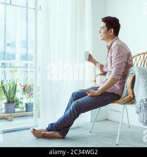 Young Asian man in cafe. Sits on aimchair at long desk near window. Stock Photo