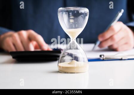 Close-up Of Hourglass In Front Of Businessperson's Hand Calculating Invoice Stock Photo
