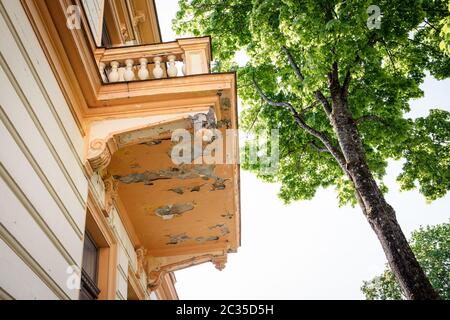 The facade of the building in poor condition. Balcony with peeling paint and cracks. Ornaments and columns Stock Photo