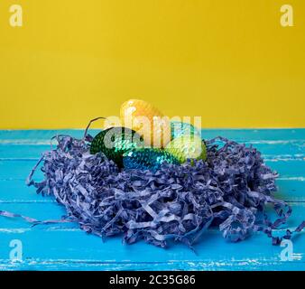 decorative Easter eggs from sequins lie in a nest of strips of paper on a colored background, copy space Stock Photo
