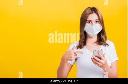 Portrait Asian Thai beautiful happy young woman wearing mask protection, pointing finger on smartphone health care, studio shot isolated on yellow bac Stock Photo