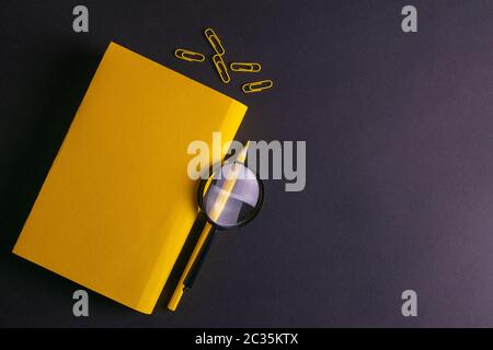 Yellow pencil, yellow notebook glasses, pins, clip paper and a magnifying glass on on black background. Stock Photo
