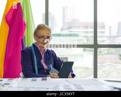Morning atmosphere in the tailor's shop. A senior designer uses her tablet computer to research information for modern design. A clothes rack with col Stock Photo