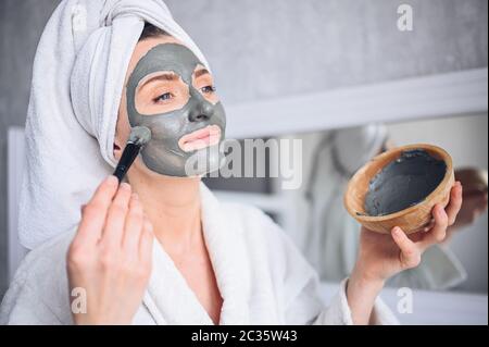 Beautiful smiling woman at home in bathrobe with a towel applying face clay mask against acne to rejuvenate problem skin Stock Photo